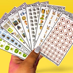 May include: A hand holds a fan of nail art decals with various designs. The decals feature images of books, dogs, and other graphics. The text "JVA NAIL SUPPLY" is visible on the decals. The background is a solid yellow color.