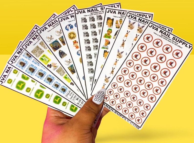 May include: A hand holds a fan of nail art decals with various designs. The decals feature images of books, dogs, and other graphics. The text "JVA NAIL SUPPLY" is visible on the decals. The background is a solid yellow color.