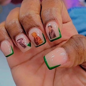 Peut inclure: Gros plan sur une main avec plusieurs ongles peints d'un vernis rose clair et de pointes vertes. Certains ongles ont des images de personnes dessus, et un ongle a le mot "Caiem" &eacute;crit dessus.