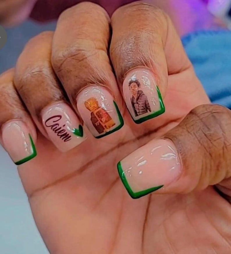 Peut inclure: Gros plan sur une main avec plusieurs ongles peints d'un vernis rose clair et de pointes vertes. Certains ongles ont des images de personnes dessus, et un ongle a le mot "Caiem" &eacute;crit dessus.