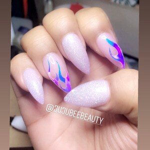 May include: White stiletto nails with iridescent flame designs. The nails are decorated with glitter. The caption on the image reads "@jujubeebeauty".