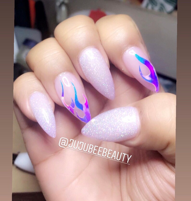 May include: White stiletto nails with iridescent flame designs. The nails are decorated with glitter. The caption on the image reads "@jujubeebeauty".