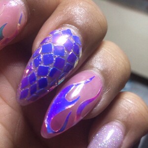 May include: Close-up of a hand with long, pointed nails. The nails are painted with a variety of designs, including a blue and purple iridescent scale pattern, a purple and pink iridescent flame pattern, and a pink and white glitter gradient.