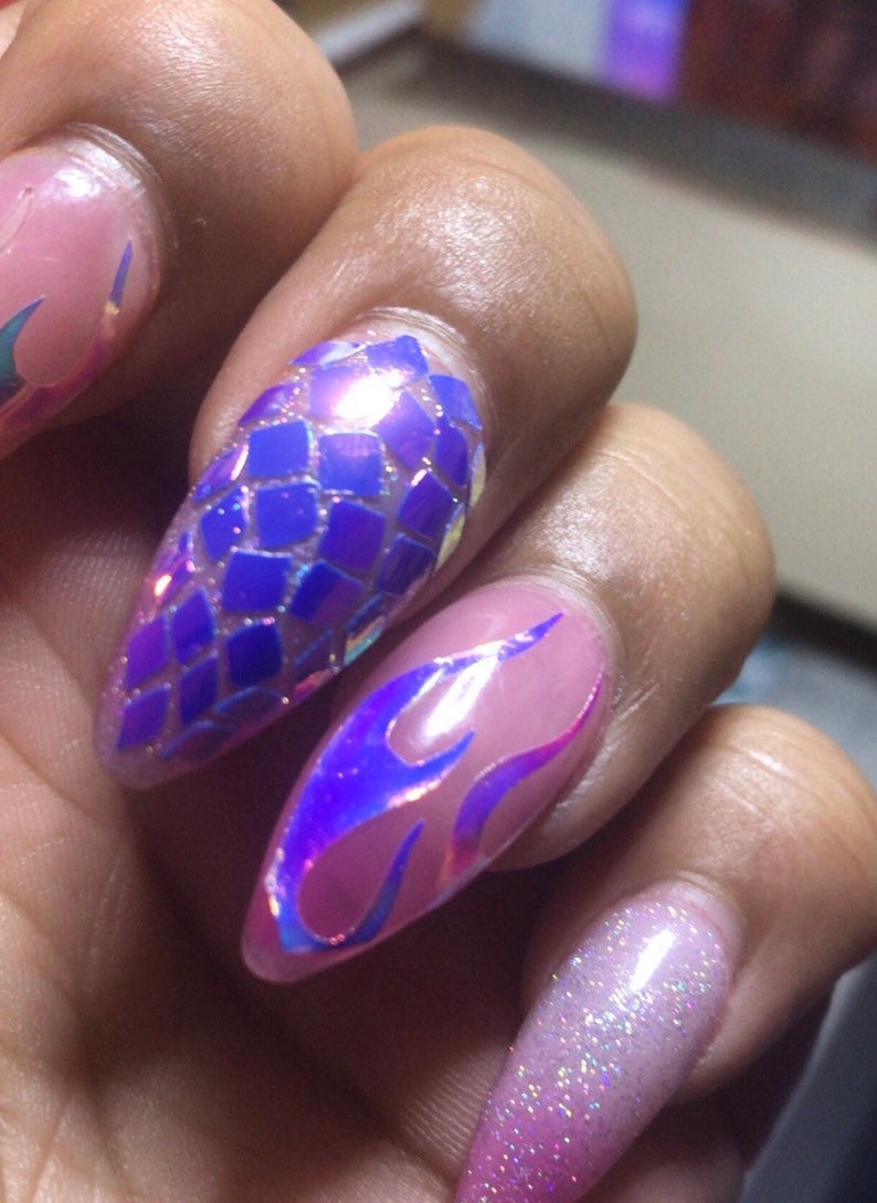 May include: Close-up of a hand with long, pointed nails. The nails are painted with a variety of designs, including a blue and purple iridescent scale pattern, a purple and pink iridescent flame pattern, and a pink and white glitter gradient.