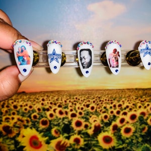 May include: White, almond-shaped artificial nails with various designs. Some nails feature images of people, while others display the Dallas Cowboys logo. Each nail is adorned with a ring of iridescent rhinestones and a blue dot at the base. The nails are displayed against a sunflower field backdrop.