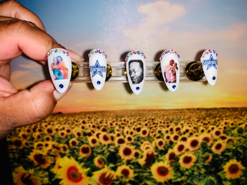 May include: White, almond-shaped artificial nails with various designs. Some nails feature images of people, while others display the Dallas Cowboys logo. Each nail is adorned with a ring of iridescent rhinestones and a blue dot at the base. The nails are displayed against a sunflower field backdrop.