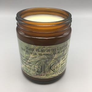 Może przedstawiać: Brązowy szklany słoik wypełniony kremowym, kremowo-białym balsamem. Słoik ma etykietę z napisem "Organic Tea Tree Oil Face & Body Balm". Etykieta zawiera również słowa "Nourishes, Softens, and Promotes Healing of All Skin". Produkt pochodzi od "JLK Body Butters".