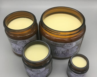 May include: Four amber glass jars of body balm with labels that read "Plumeria Organic Body Balm" and "Nourishes and softens skin of all types". The jars are filled with a pale yellow balm and are arranged in a pyramid shape.