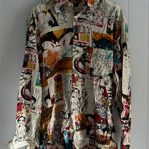 Pode incluir: Uma camisa de manga comprida com botões, fundo branco e estampa abstrata colorida. A estampa apresenta várias figuras, texto e formas geométricas. A camisa tem um caimento solto e gola.