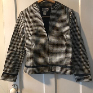 Puede incluir: Una chaqueta de punto de pata de gallo en blanco y negro con ribete negro alrededor del cuello, las mangas y el dobladillo. La chaqueta tiene cuello en V y cierre de cremallera. La etiqueta de la chaqueta dice "Pendleton".