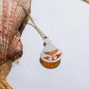 May include: A silver-toned pendant necklace featuring a cutout design of a whale tail. The pendant has a translucent orange insert and hangs from a delicate silver-toned chain. The piece is positioned next to a large, textured seashell.
