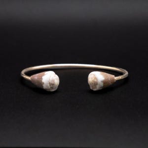 Può includere: Un bracciale rigido in metallo color oro dal design aperto, caratterizzato da due piccole conchiglie coniche marroni e bianche fissate alle estremità. Il cerchio è sottile e liscio, rimanendo aperto sulla parte anteriore per facilitare l'uso.