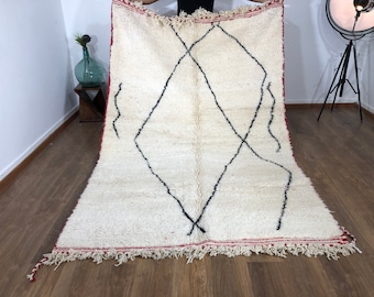 May include: A white Moroccan rug with a black diamond pattern and red fringe. The rug is laid out on a wooden floor.