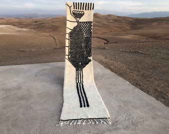 May include: A rectangular, off-white rug with a black geometric design. The design includes vertical lines, a checkered pattern, and abstract shapes. The rug has a fringe at the bottom and is displayed outdoors.
