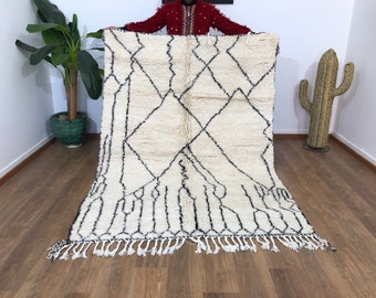 May include: A white Moroccan rug with a black diamond pattern. The rug has a fringe border and is laid out on a wooden floor.