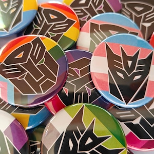 May include: A collection of circular pin-back buttons featuring the black Decepticon logo centered on various backgrounds, including rainbow stripes, pink, white, and blue horizontal stripes, and other solid color patterns with a glossy finish.