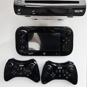 May include: A black Wii U console, gamepad, and two controllers are shown on a white surface. The console has a disc slot and the gamepad has a large screen. The controllers have buttons and joysticks.