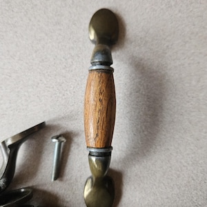 May include: A close-up of a cabinet handle with a wooden centre and brass-coloured ends. The handle is accompanied by a screw and other hardware. The wood grain is visible, and the metal has a slightly aged appearance.