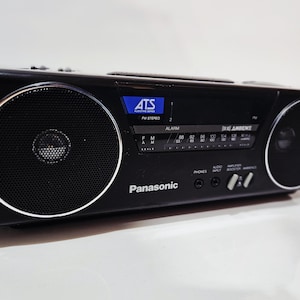 May include: Black Panasonic AM/FM stereo radio with two circular speakers and a digital display. The radio has a silver trim around the speakers and a series of buttons and dials. The text "ATS" and "FM STEREO" are visible on the display.