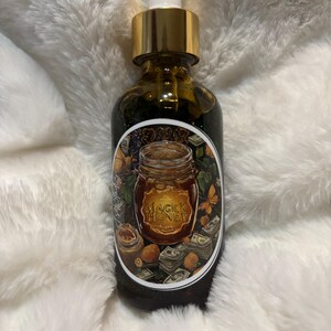 May include: A brown glass bottle with a gold cap and white dropper. The bottle has a label with the words "Magick Honey" and a jar illustration. The bottle is filled with a dark liquid and sits on a white, fluffy surface.