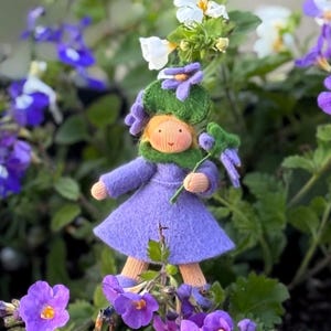 Könnte beinhalten: Eine lila Filzpuppe mit einem grünen Filzhut und einer lila Blume in der Hand. Die Puppe steht in einem Beet mit lila Blumen.