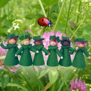 Puede incluir: Imagen caprichosa de seis muñecas de fieltro con vestidos y sombreros verdes, tomadas de la mano, en medio de flores. Una mariquita roja descansa encima. Las muñecas tienen diferentes tonos de piel y colores de cabello.
