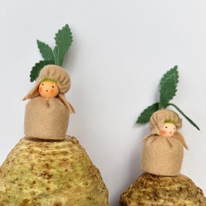 May include: Two whimsical handmade dolls, each featuring a tan felt body and a small doll head. The dolls are perched atop large, textured root vegetables, with green felt leaves sprouting from the top. The background is a plain white wall.
