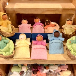 Puede incluir: Una colecci&oacute;n de mu&ntilde;ecas de fieltro hechas a mano en varios colores, cada una acostada en una cama de fieltro o una peque&ntilde;a cuna de madera. Las mu&ntilde;ecas est&aacute;n vestidas con atuendos con capucha y tienen caras pintadas.