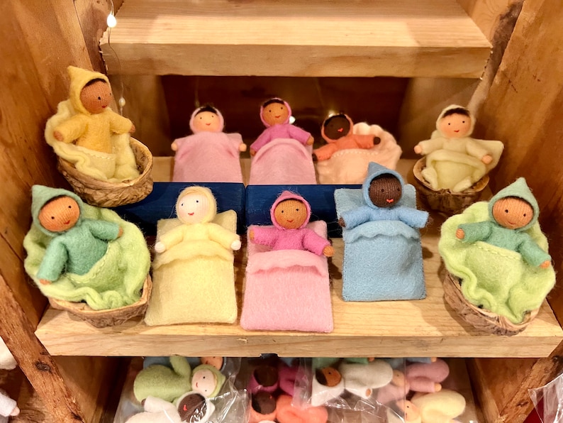 Puede incluir: Una colecci&oacute;n de mu&ntilde;ecas de fieltro hechas a mano en varios colores, cada una acostada en una cama de fieltro o una peque&ntilde;a cuna de madera. Las mu&ntilde;ecas est&aacute;n vestidas con atuendos con capucha y tienen caras pintadas.