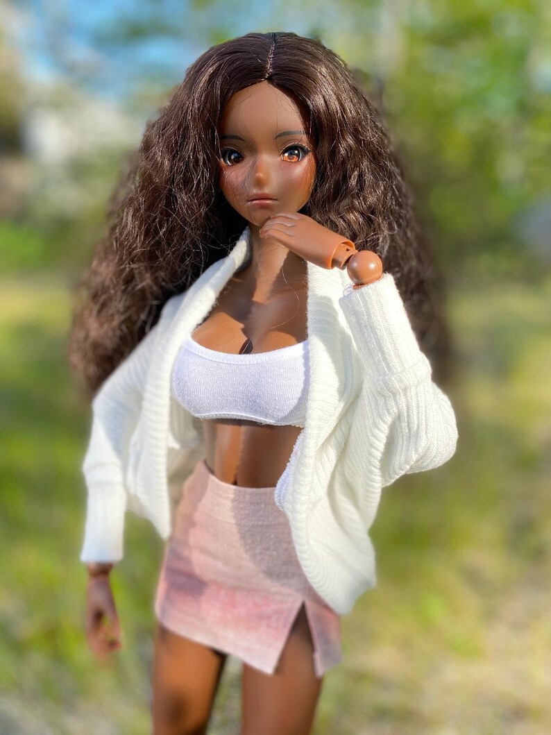 Op de afbeelding: Een pop met lang bruin krullend haar, die een witte cardigan, een wit crop top en een roze rok met een split draagt.