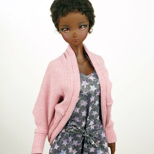 Op de afbeelding: Een pop met donkerbruin krullend haar die een roze cardigan draagt over een top met sterrenprint en een bijpassende short.