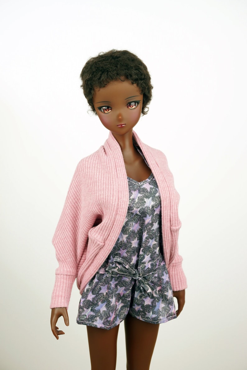 Op de afbeelding: Een pop met donkerbruin krullend haar die een roze cardigan draagt over een top met sterrenprint en een bijpassende short.