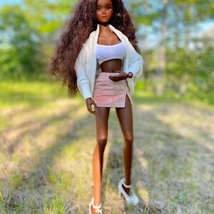 Op de afbeelding: Een modepop met lang, bruin, krullend haar, die een witte cardigan, een roze rok en witte schoenen draagt. De pop staat in een grasveld.