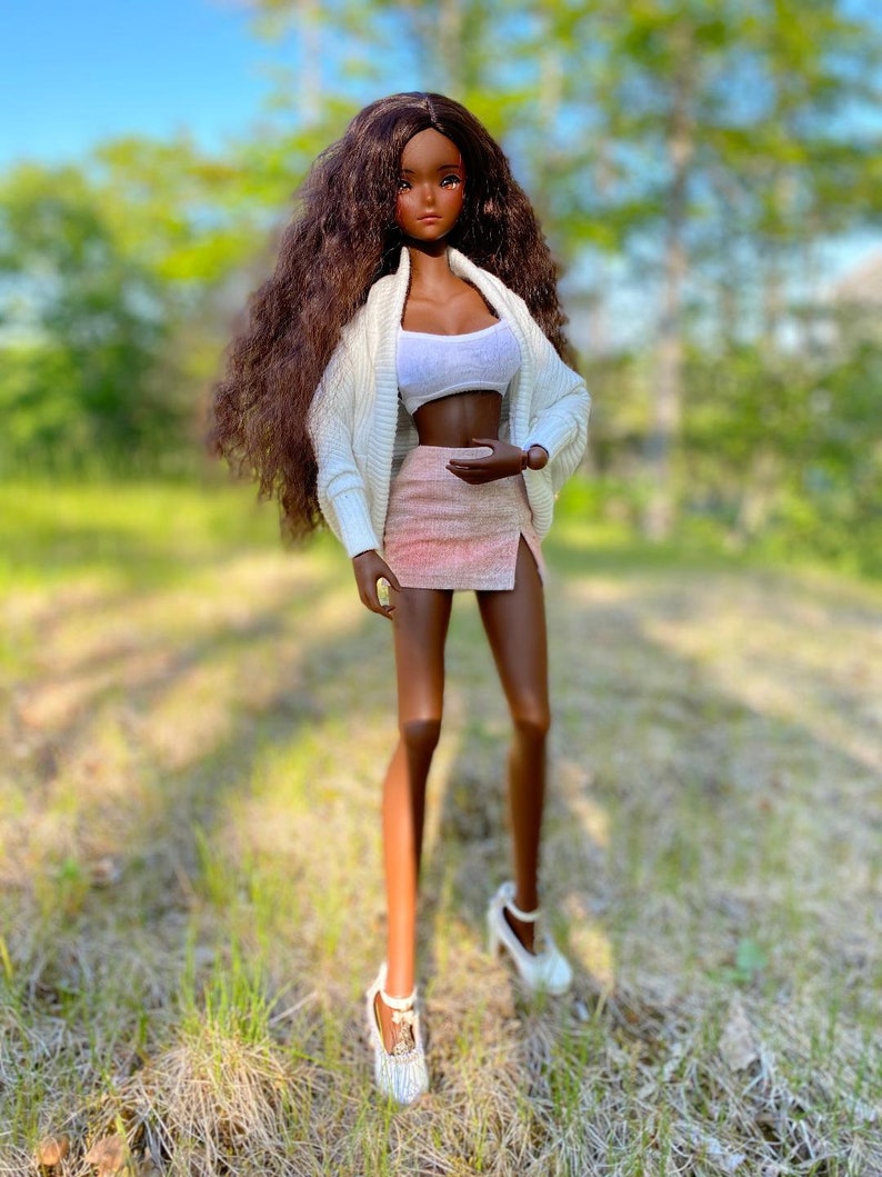 Op de afbeelding: Een modepop met lang, bruin, krullend haar, die een witte cardigan, een roze rok en witte schoenen draagt. De pop staat in een grasveld.