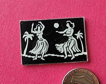 May include: A black and white enamel pin featuring two figures in hula skirts dancing with a palm tree on each side. The pin is approximately the size of a US penny.