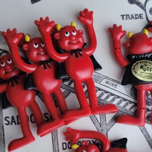 May include: Five red plastic devil figurines with black capes and yellow horns. The figurines have bendable arms and legs. The figurines are on a white surface with black and white text.