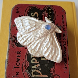 May include: A pearlescent moth-shaped brooch with a blue gemstone eye rests on a vintage red and black tin. The tin features the text "The Tower" and "Paper Box". The moth has detailed wing patterns.