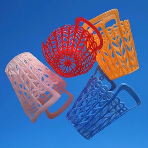 May include: Four brightly coloured, openwork baskets in pink, red, orange, and blue against a blue background. Each basket has a handle and a unique geometric or floral design, showcasing a variety of textures and patterns.