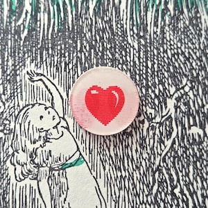 May include: A round, clear button with a red pixelated heart design. The button is set against a black and white illustration of a girl and a dog, with green and black foliage in the background.