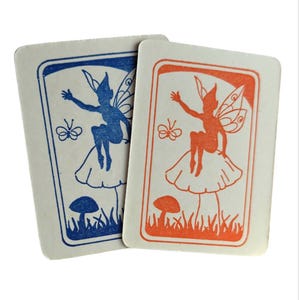 May include: Two cards with a fairy sitting on a mushroom with butterflies. One card is blue and the other is orange.