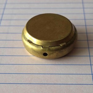 May include: A small, circular, brass-colored metal container with a smooth, flat top and a rounded side. The side features a single, small, dark circular hole and a visible hinge mechanism connecting the lid to the base of the object.