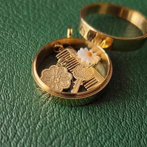 May include: A gold-toned circular locket pendant with a detachable ring top, containing small charms including a gold comb, a gold key, a white daisy flower with a yellow center, and a gold flower-shaped charm embossed with the number 13.