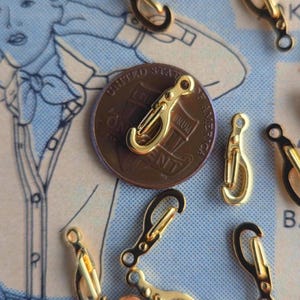 May include: A collection of small gold-toned metal lobster claw clasps scattered around a copper one-cent coin. Each clasp features a spring-loaded gate and a small circular loop at the base for attachment, shown against a blue patterned background.