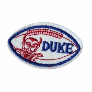 May include: Embroidered patch in an oval shape with a white background. The patch features a red devil with horns and the word "DUKE" in blue. A blue and red border surrounds the design.