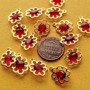 Puede incluir: Una colección de 13 abalorios de metal dorado con piedras de vidrio rojo en el centro. Cada abalorio tiene forma de flor con un bucle en la parte superior para colgar.