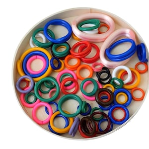 May include: A collection of colorful rings in various sizes and shades, including blue, green, pink, red, orange, yellow, and brown. The rings are arranged inside a round, light-colored wooden tray. The image is well-lit, showcasing the vibrant colors and textures of the rings.