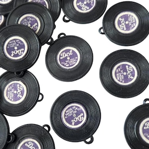 May include: A collection of black circular plastic charms shaped like miniature vinyl records. Each charm features a central purple label with the text Dot, I'll See You In My Dreams, and Pat Boone, with a small loop at the top for attachment.