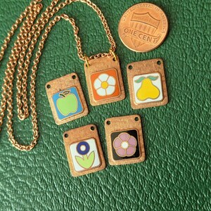 May include: A copper-toned metal necklace with a chain and five rectangular pendants featuring enamel designs of a green apple, a white flower on orange, a yellow pear, a blue flower on white, and a pink flower on black, next to a US one cent coin.