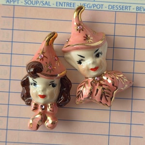 May include: Two vintage ceramic figurines of women's faces. They have pink hats with gold trim and star accents, and pink clothing with gold details. The faces have red lips and dark hair.