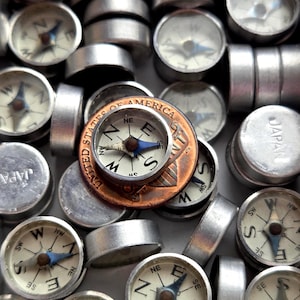 May include: A collection of small, silver-colored compasses with white faces and blue needles. A copper penny with the words "United States of America" is in the center. The compasses are arranged closely together.
